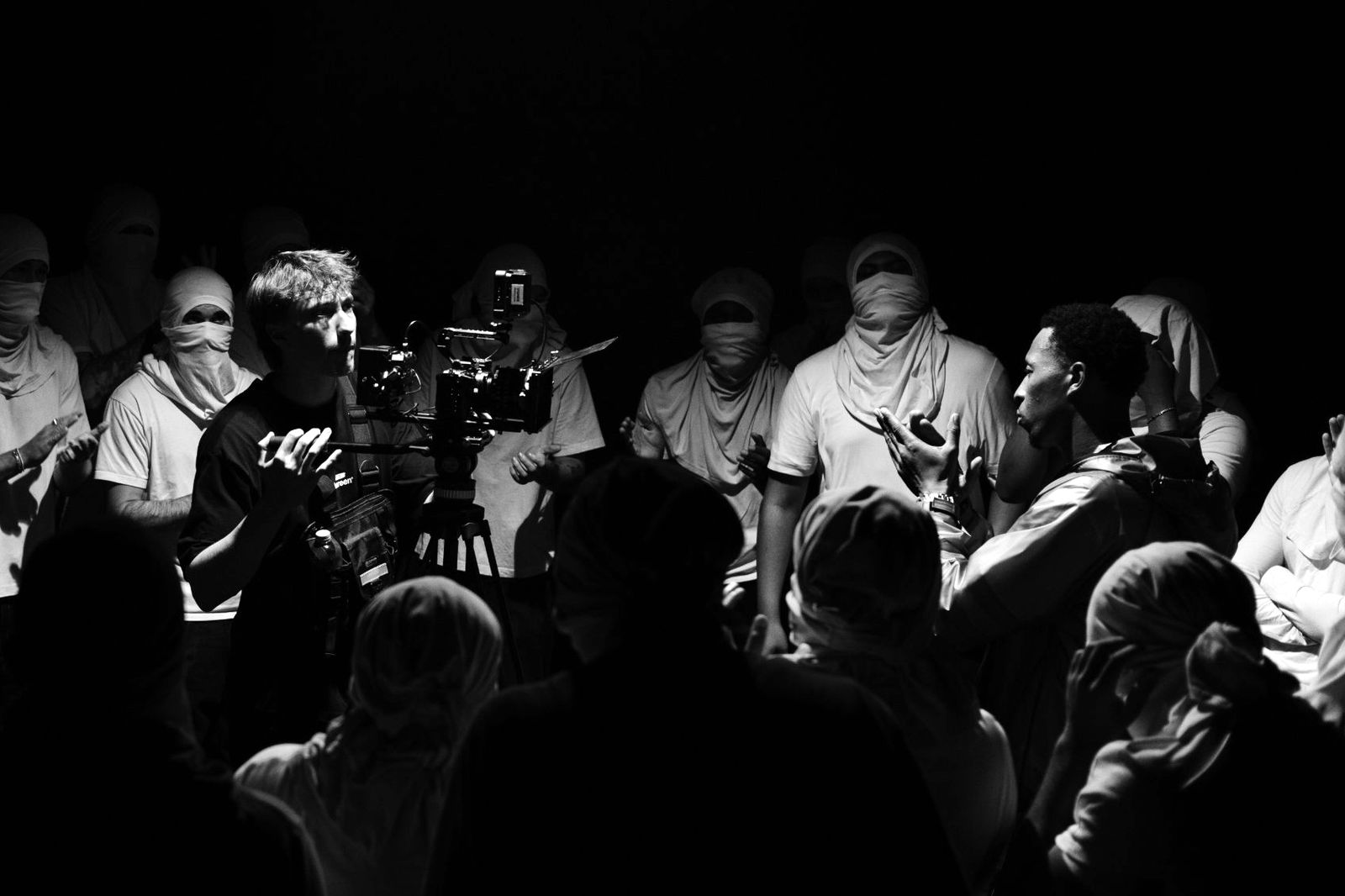 Oliver filming with a camera on a tripod next to the artist, surrounded by a crowd in white face coverings