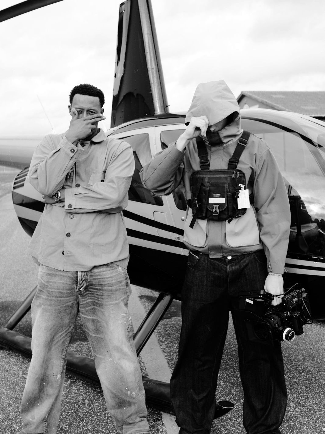 Oliver in a hooded jacket holding a cinema camera next to an artist in front of a helicopter