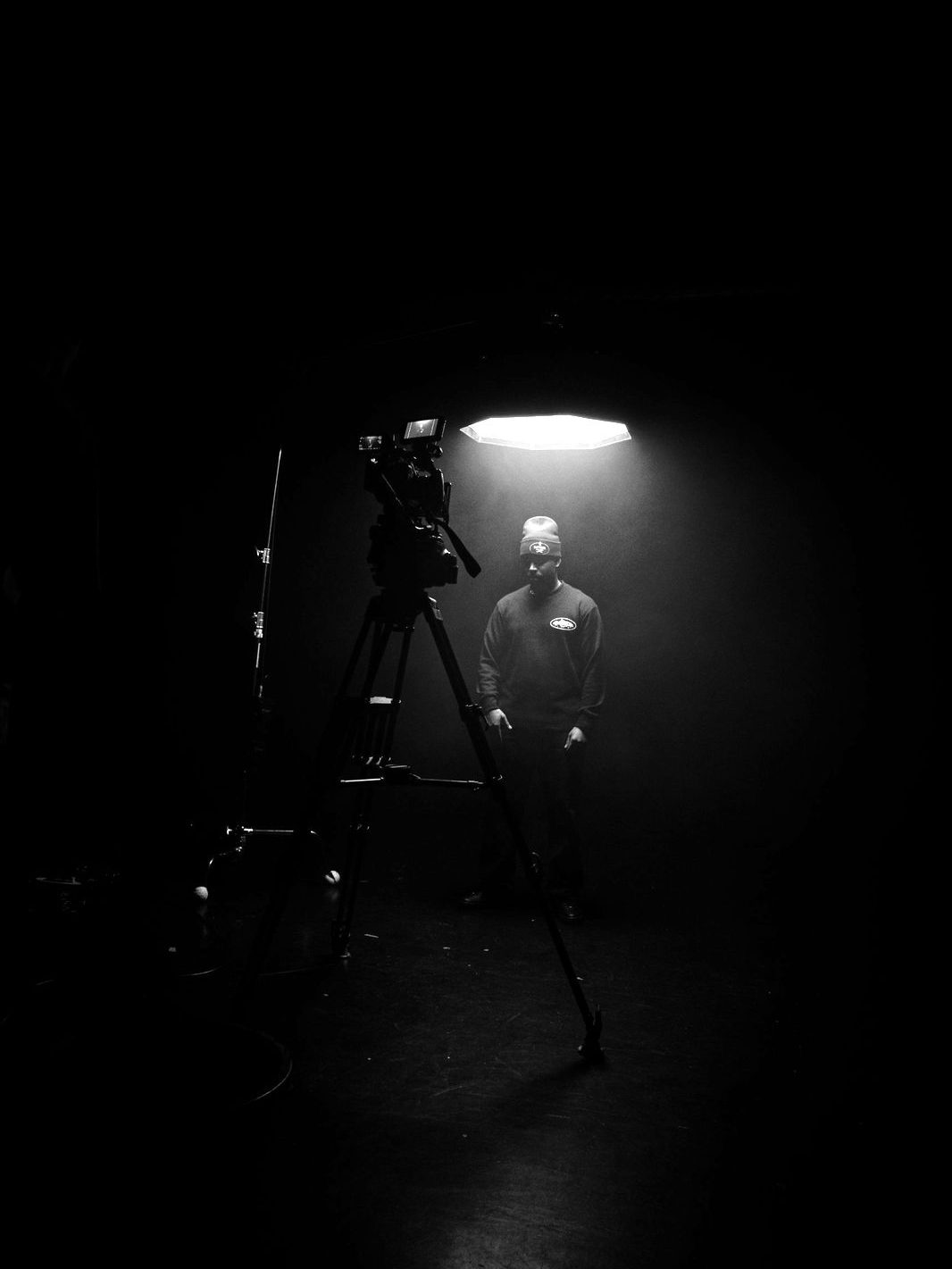 A camera on a tripod facing an artist standing under a single overhead light
