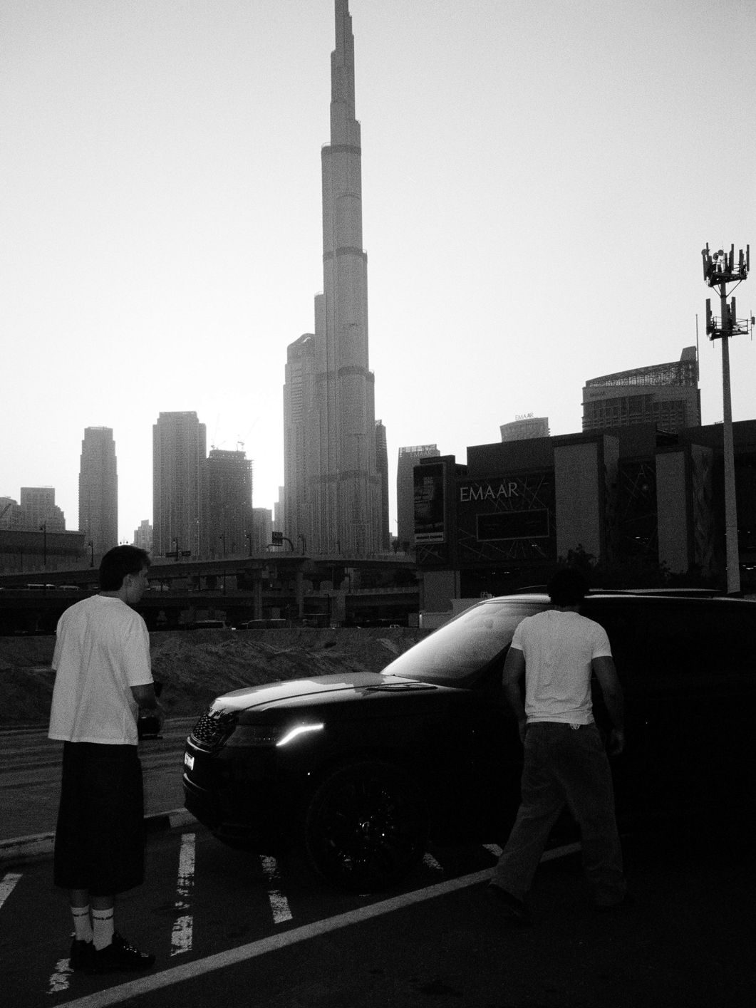 Two crew members by a black car in a Dubai parking lot, with the Burj Khalifa behind them at sunset